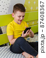 The boy plays on a smartphone while sitting in his room on the bed. 87408367