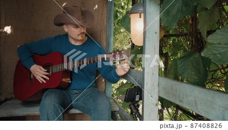 Man with guitar in a cowboy hat sits on the porch and sings songs while reading the text from his phone. The concept of recreation, hobbies and lifestyle. Old, retro decor, burlap background Man with guitar in a cowboy hat sits on the porch and sings songs while reading the text from his phone. The concept of recreation, hobbies and lifestyle. Old, retro decor, burlap background 87408826