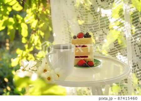 Beautiful porcelain coffee cup and slice of cake on glass table decorated with white flowers in summer garden in sunlight. Copy space. Summer drinks concept. Beautiful porcelain coffee cup and slice of cake on glass table decorated with white flowers in summer garden in sunlight. Copy space. Summer drinks concept. 87415460