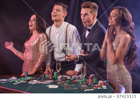 Friends playing poker at casino, at table with stacks of chips, money, cards, champagne on it. Celebrating win, excited. Black background. Close-up. Friends playing poker at casino, at table with stacks of chips, money, cards, champagne on it. Celebrating win, excited. Black background. Close-up. 87423115