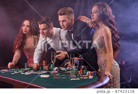 Friends playing poker at casino, at table with stacks of chips, money, cards, champagne on it. Celebrating win, excited. Black background. Close-up. Friends playing poker at casino, at table with stacks of chips, money, cards, champagne on it. Celebrating win, excited. Black background. Close-up. 87423116