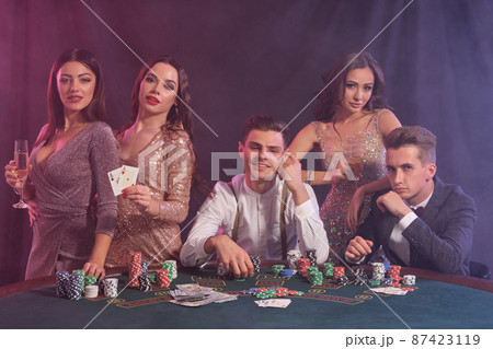 Friends playing poker at casino, at table with stacks of chips, money, cards on it. Celebrating win, smiling. Black, smoke background. Close-up. Friends playing poker at casino, at table with stacks of chips, money, cards on it. Celebrating win, smiling. Black, smoke background. Close-up. 87423119