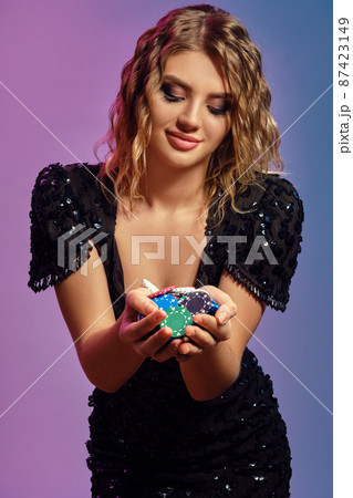 Blonde girl in black sequin dress is showing handful of multicolored chips, posing on colorful studio background. Gambling, poker, casino. Close-up. 87423149