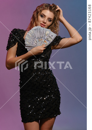 Brown-haired girl in black shiny dress is holding some cash, posing against colorful background. Gambling entertainment, poker, casino. Close-up. 87423168