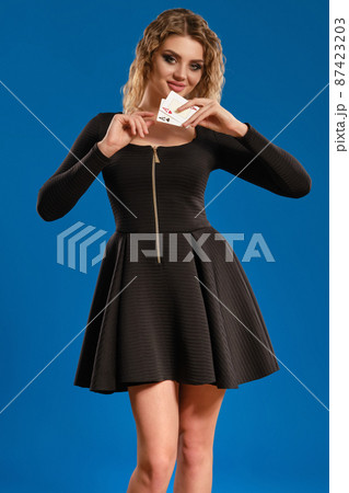 Blonde girl in black short dress is smiling, showing two aces, posing against blue background. Gambling entertainment, poker, casino. Close-up. Blonde girl in black short dress is smiling, showing two aces, posing against blue background. Gambling entertainment, poker, casino. Close-up. 87423203
