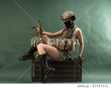 a girl soldier armed with an automatic rifle in military clothes sits on a gun case on a green background 87437331