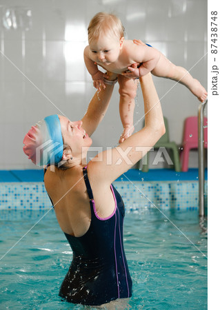 Early age swimming in pool. Baby boy trained to swim in water. Happy child with trainer woman in indoor swimming pool playing and having fun. Healthy and sport family with infant, active parent Early age swimming in pool. Baby boy trained to swim in water. Happy child with trainer woman in indoor swimming pool playing and having fun. Healthy and sport family with infant, active parent 87438748
