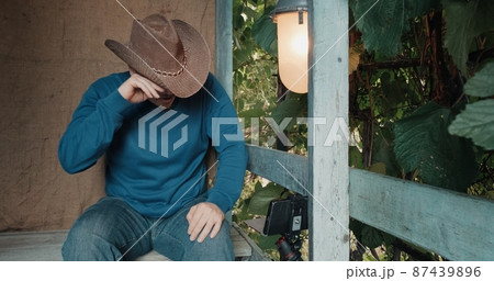 Man in a cowboy hat is sitting on the porch of an old house and looking at the phone, something very funny. The man bursts into laughter. Concept of humor and parody 87439896