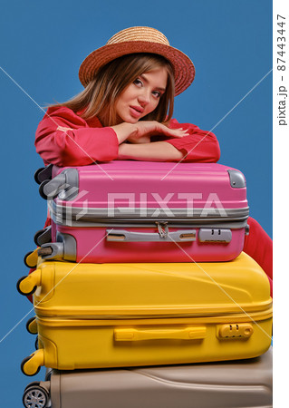 Blonde lady in straw hat and red jacket. Sitting leaning on colorful suitcases, posing on blue background. Travelling concept. Close-up 87443447