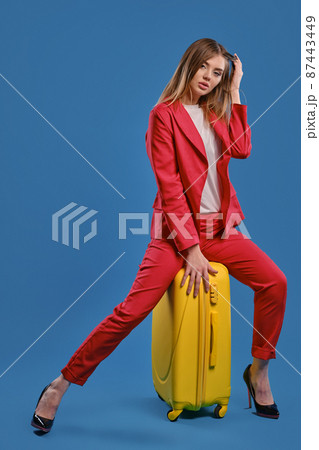 Blonde lady in white blouse, red pantsuit, high black heels. She touching hair, sitting on yellow suitcase, posing on blue background. Full length Blonde lady in white blouse, red pantsuit, high black heels. She touching hair, sitting on yellow suitcase, posing on blue background. Full length 87443449