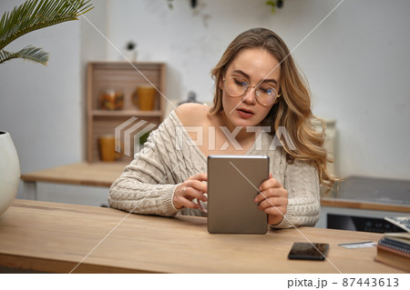 Girl blogger in glasses, beige sweater. Holding tablet, sitting in kitchen at table with notebooks, smartphone, plastic card and palm on it. Close-up 87443613