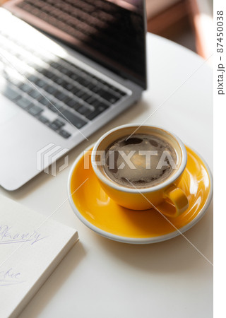 Yellow cup of coffee on workspace on table near laptop.  87450038