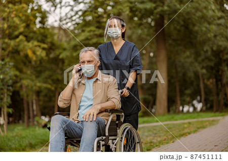 Young nurse in face shield and protective face mask taking care of senior handicapped patient in wheelchair talking on the phone during a walk in the park 87451111