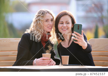 Two happy women making selfie with mobile phone sitting at city street cafe. Female friends enjoying time together outdoors Two happy women making selfie with mobile phone sitting at city street cafe. Female friends enjoying time together outdoors 87452337