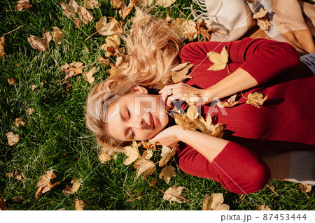 an attractive blonde woman lies on a green lawn with autumn leaves.  87453444