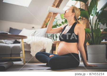 Young happy and cheerful beautiful pregnant woman taking a break, hydrating, drinking water from the botle after home well being workout progrem. 87455254