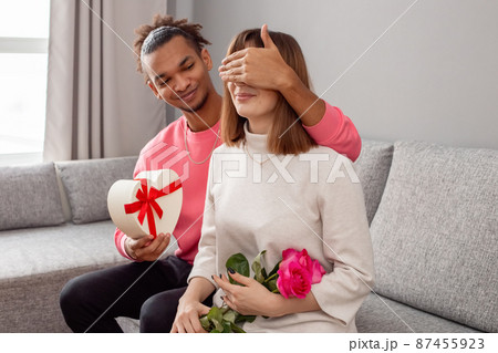 A guy give a surprise at a girl , sits on a gray sofa. 87455923