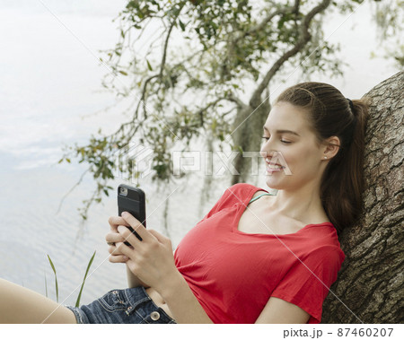 Smiling woman leaning on tree branch and looking at smart phone 87460207