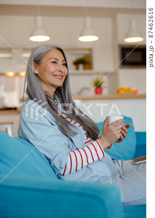 Cheerful woman with cup of coffee sitting on couch at home Cheerful woman with cup of coffee sitting on couch at home 87462616