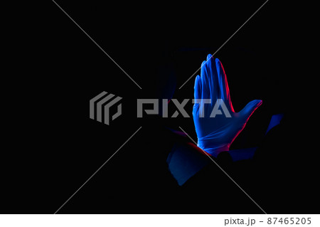 Doctor warning. Stop gesture. Red blue neon light hand in glove showing restriction palm in black breakthrough torn wall hole isolated on dark night empty space. Doctor warning. Stop gesture. Red blue neon light hand in glove showing restriction palm in black breakthrough torn wall hole isolated on dark night empty space. 87465205