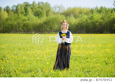 Young woman in national clothes wearing yellow dandelion wreath in spring field. Springtime Young woman in national clothes wearing yellow dandelion wreath in spring field. Springtime 87470451