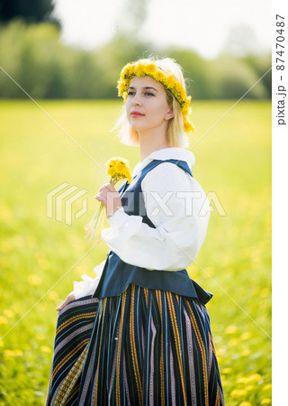 Young woman in national clothes wearing yellow dandelion wreath in spring field. Springtime Young woman in national clothes wearing yellow dandelion wreath in spring field. Springtime 87470487