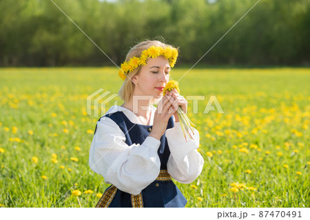 Young woman in national clothes wearing yellow dandelion wreath in spring field. Springtime Young woman in national clothes wearing yellow dandelion wreath in spring field. Springtime 87470491