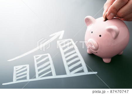 将来に備え計画的に貯金をするイメージ。ブタの貯金箱にコインを入れる。黒板に描かれたグラフと矢印。 87470747