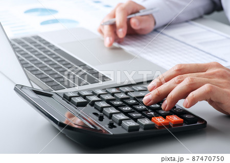 会計と計算のイメージ。電卓で計算をする手のアップ。机の上にノートパソコンとレポート。 87470750
