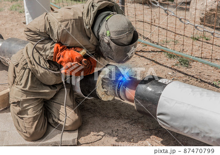 A welder in a protective shield is engaged in welding work on the polyethylene pipe of the heating main pipeline at the construction site 87470799