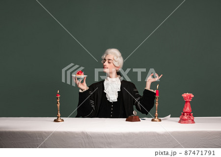 Portrait of young elegant man in peruke and vintage jacket sitting at table isolated on dark green background. Retro style, comparison of eras concept. 87471791