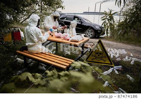 Environmentalists in radiation suits drinking in roadside forest with plastic bottles and poison toxic sign. Family resting at polluted territory with garbage and skull-and-crossbones warning symbol. 87474186