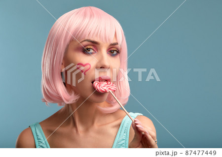 Close up of a flirtatious female model with a candy in mouth. Pretty glamorous woman pink hair charm sweets lifestyle enjoyment sugar 87477449