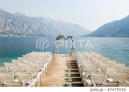 Wedding arch stands in front of a row of chairs on a wooden pier overlooking the mountains 87479550