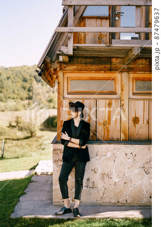 Young man in a hat stands near an old wooden house with his arms folded on his chest 87479637