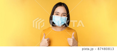Cheerful korean woman in medical face mask, support people during pandemic, wear personal protective equipment from covid-19, showing thumbs up in approval, yellow background 87480813