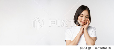 Portrait of tender and feminine young asian woman, looking down, gently touching face, concept of skin care and cosmetology, white background 87480816