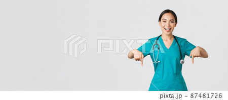 Covid-19, healthcare workers, pandemic concept. Happy smiling beautiful asian nurse, female doctor in scrubs pointing down, inviting for checkup, showing hospital advertisement, white background 87481726