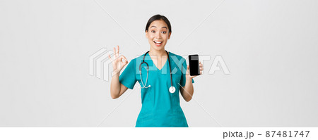 Covid-19, healthcare workers and online medicine concept. Impressed and pleased asian female doctor trying new online consultation method during coronavirus, showing smartphone app and okay gesture Covid-19, healthcare workers and online medicine concept. Impressed and pleased asian female doctor trying new online consultation method during coronavirus, showing smartphone app and okay gesture 87481747