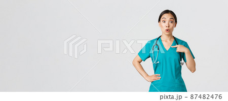 Covid-19, healthcare workers, pandemic concept. Surprised and ambushed asian female physician, nurse in scrubs pointing at herself, beaing named or chosen, standing white background 87482476