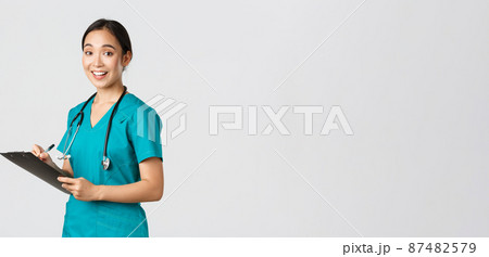 Healthcare workers, preventing virus, quarantine campaign concept. Smiling cute asian female nurse, intern in scrubs, writing on clipboard, fill-in papers, looking a camera pleased, white background 87482579