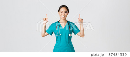 Covid-19, healthcare workers, pandemic concept. Happy smiling asian female doctor, nurse in scrubs showing advertisement, making announcement. Physician pointing fingers up at banner 87484539