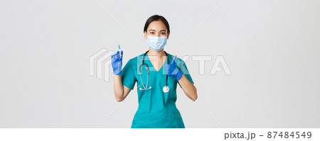 Covid-19, coronavirus disease, healthcare workers concept. Pleasant smiling asian female doctor, physician in medical mask and gloves, show thumb-up, hold syringe with vaccine 87484549