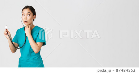 Covid-19, healthcare workers and preventing virus concept. Intrigued asian female doctor, physician in scrubs, looking away thoughtful, listening to patient lungs with stethoscope, white background 87484552