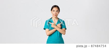 Covid-19, healthcare workers, pandemic concept. Creative and thoughtful pretty asian female nurse, intern have idea, making choice, pointing fingers sideways but looking left, thinking 87484754