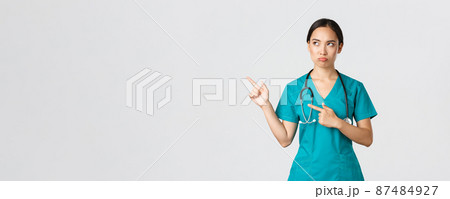 Covid-19, healthcare workers, pandemic concept. Disappointed gloomy, sulking asian nurse in scrubs, female doctor looking displeased, pointing upper left corner and frowning judgemental 87484927