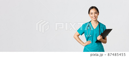 Healthcare workers, preventing virus, quarantine campaign concept. Smiling pleasant asian female physician, doctor during examination wearing scrubs and holding clipboard, white background 87485455