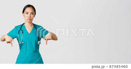 Covid-19, healthcare workers, pandemic concept. Upset and disappointed pouting asian female therapist, nurse in scrubs pointing fingers down, sulking sad, showing something with regret 87485460