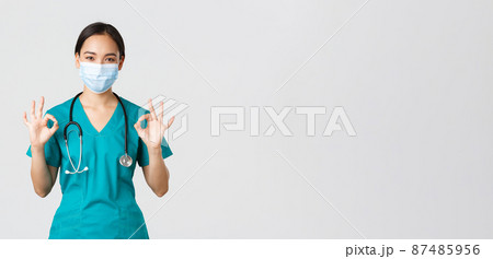 Covid-19, coronavirus disease, healthcare workers concept. Young professional female doctor, asian intern in scrubs and medical mask showing okay gesture, guarantee quality Covid-19, coronavirus disease, healthcare workers concept. Young professional female doctor, asian intern in scrubs and medical mask showing okay gesture, guarantee quality 87485956