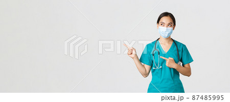 Covid-19, coronavirus disease, healthcare workers concept. Curious pleased asian female doctor, physician in medical mask and gloves, looking and pointing upper left corner with intrigued smile 87485995
